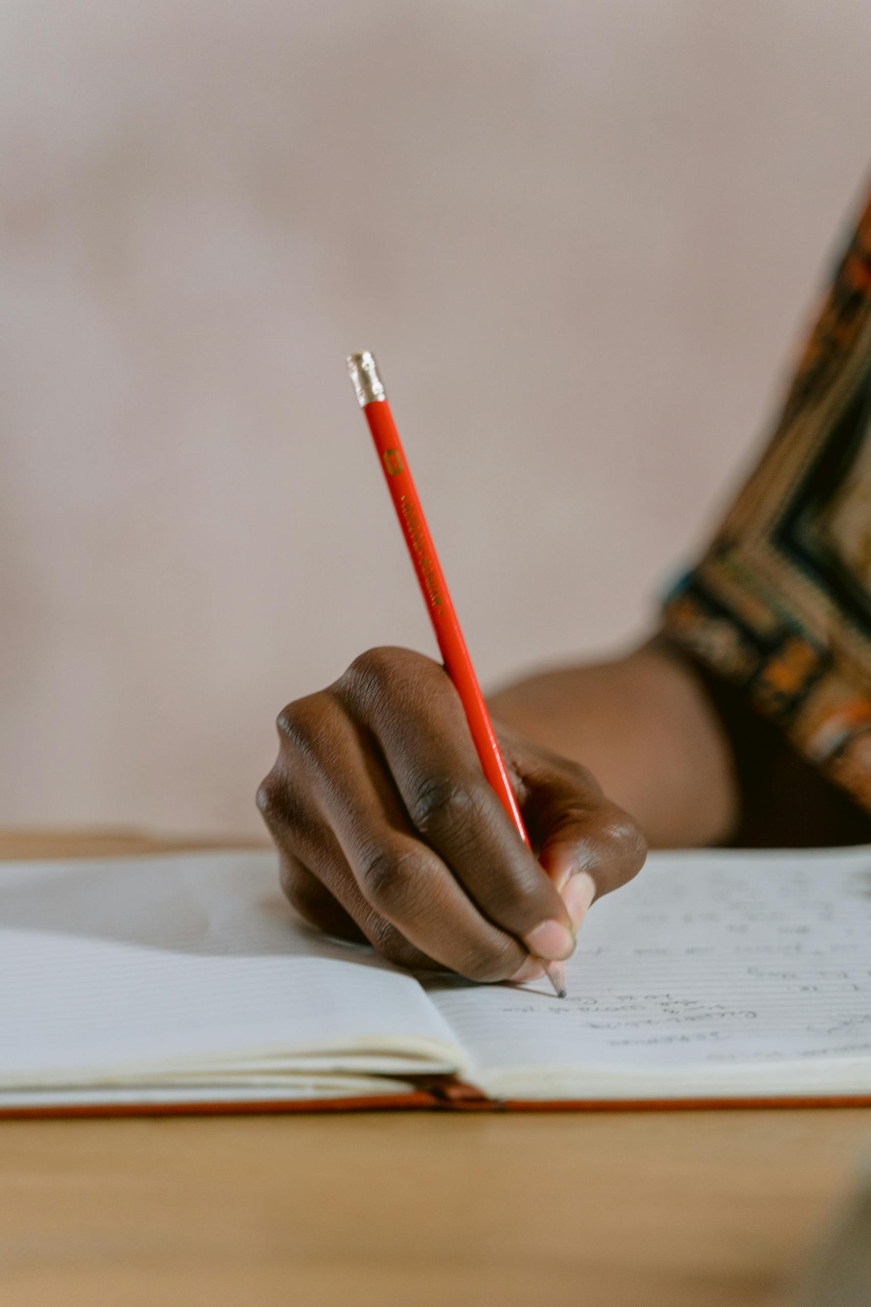 A person writing in a notebook with a red pencil, focusing on the hand's detail.